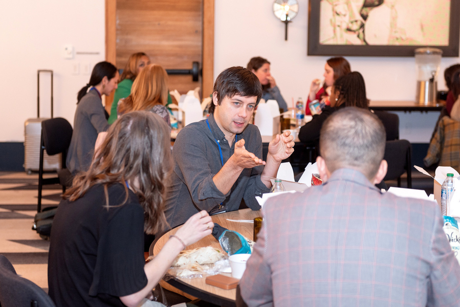 Eight people sit around two round tables, eating lunch. The only individual in focus is speaking and gesturing with his hands.