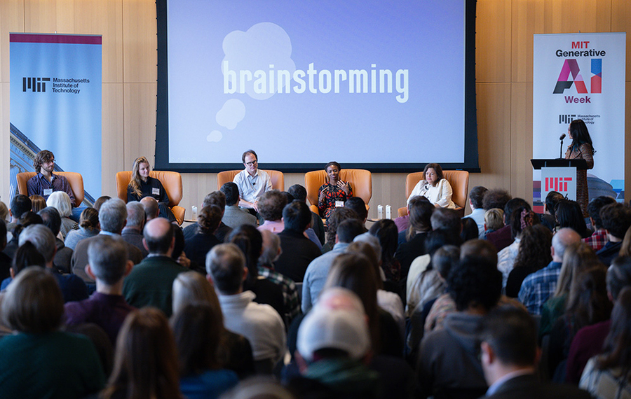 Audience watches panel of five seated individuals on stage in front of screen that reads