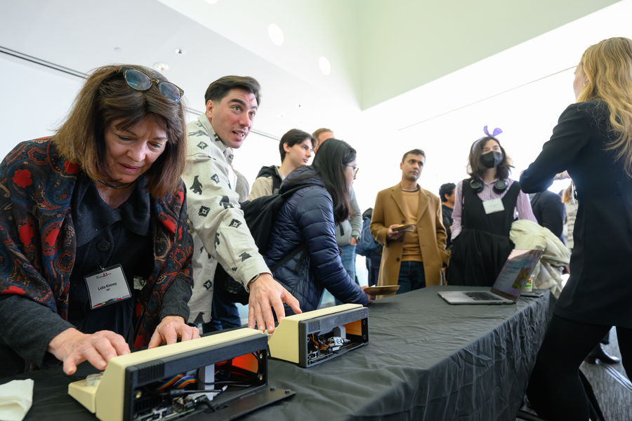 Man and woman interact with keyboard devices on a table, while others in the room mill about