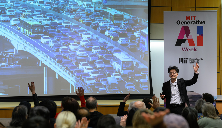 Jinhua Zhao, on stage, speaks while standing next to a screen with a photo of cars stuck in traffic