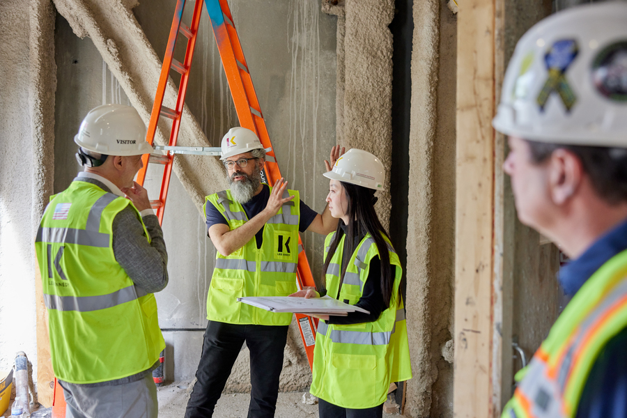 Three people wearing hardhats and fluorescent safety vests stand in a room under construction. A fourth person, looking away from the camera, is out of focus in the foreground.