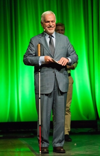 Paul Parravano stands on a stage with a green-lit curtain behind him. He is holding a paper while his white cane rests under his arm.