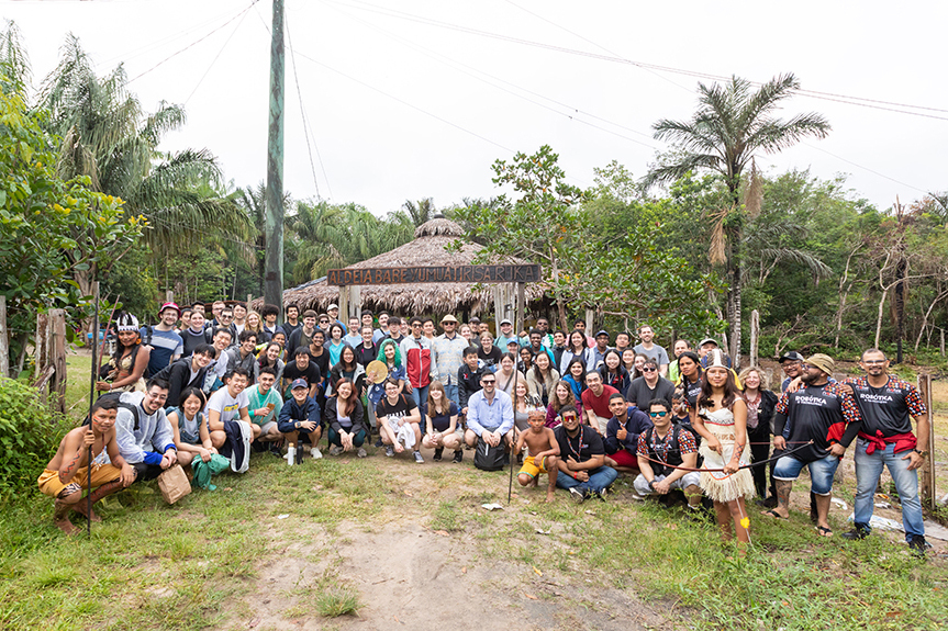 A large group of perhaps 80 MIT musicians and tour staff pose with members of the São Sebastião community in native garb with lush tropical trees in the background
