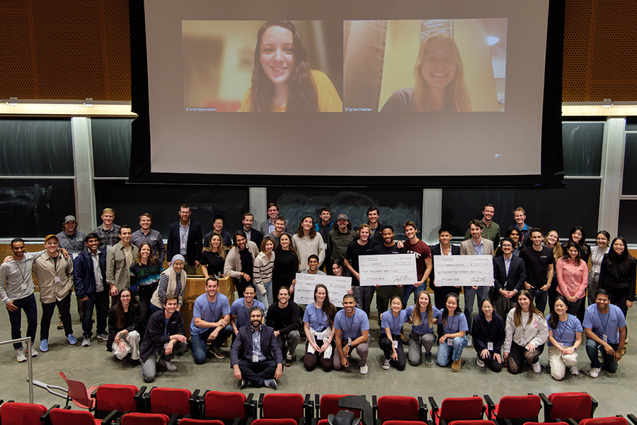 Approximately 40 people pose on a stage, under a huge screen displaying two portraits