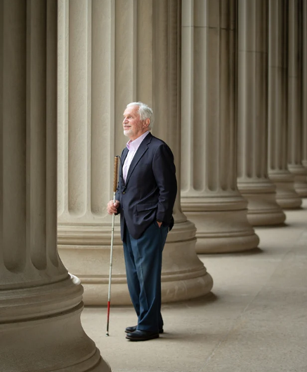 Paul Parravano stands among the columns of an iconic building on MIT’s campus, looking into the distance. He’s holding his white cane.