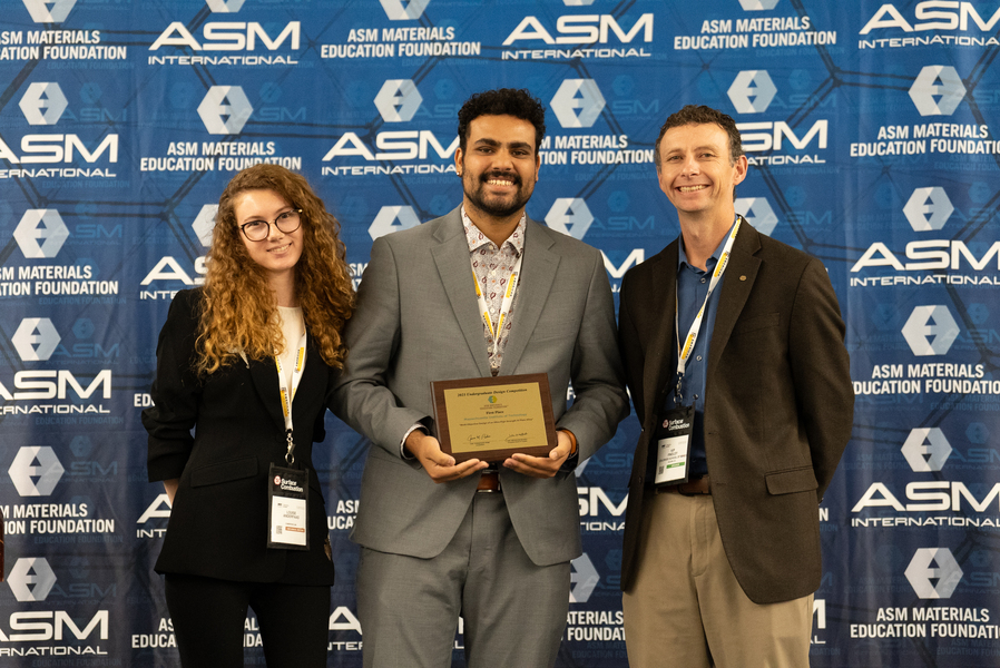 Three people stand on a stage in front of an ASM background. The one in the center holds a plaque.