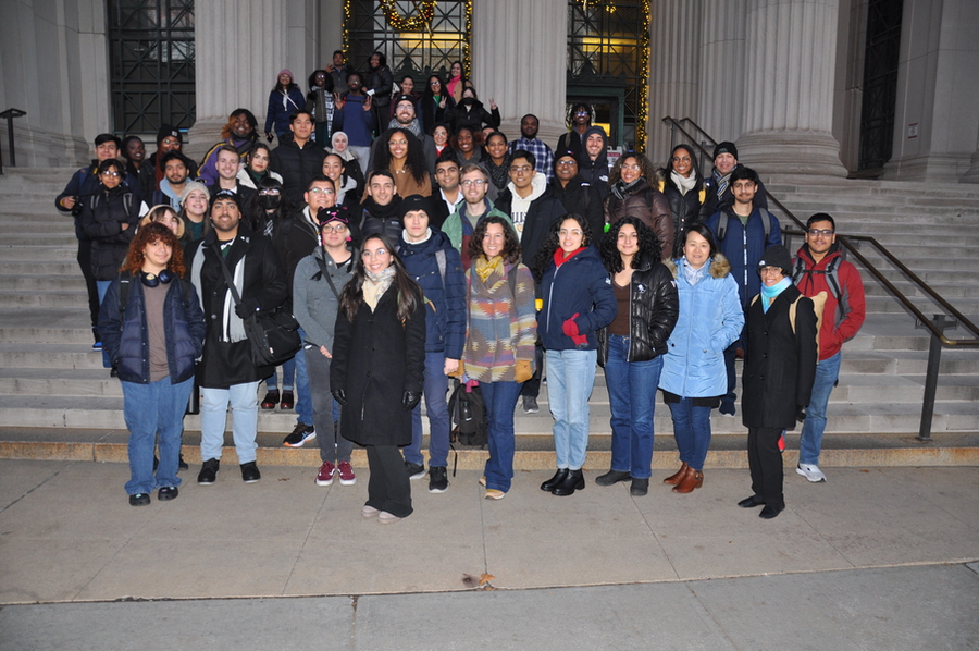 Clad in winter coats and hats, a large group of students and professors smile as they gather on the steps of MIT's main entrance at night
