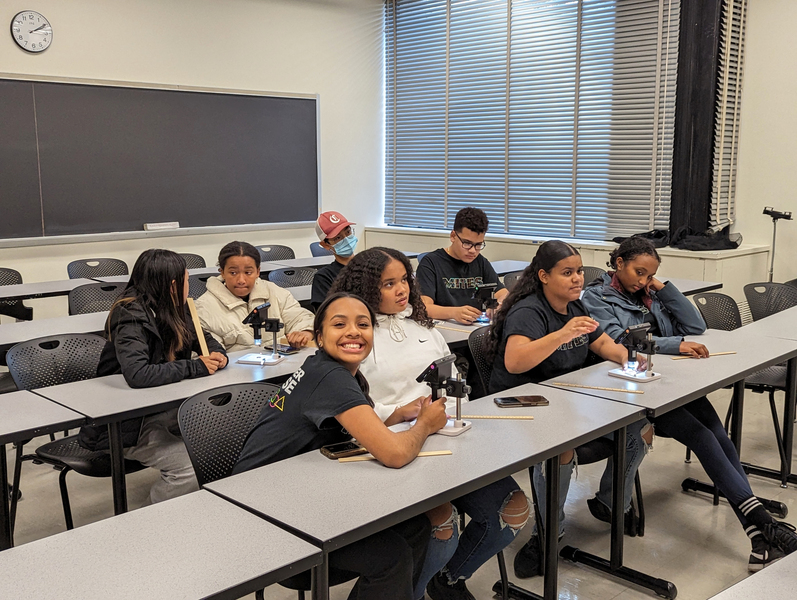 Eight middle school students sit at tables in a classroom at MIT. One is smiling at the camera.