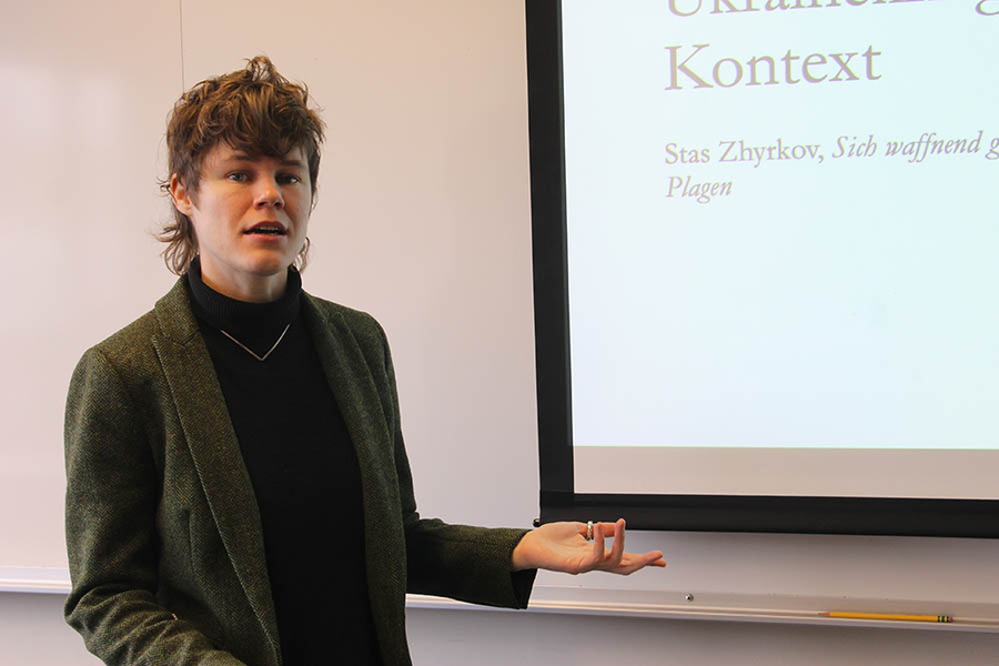 Emily Goodling stands in front of a classroom next to a Powerpoint slide with German text.