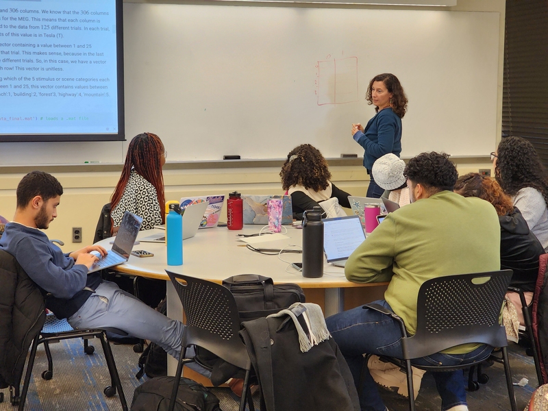 Tiziana Ligorio stands at a whiteboard next to a projector screen and looks back toward a round table of seven students who are sitting with laptops.