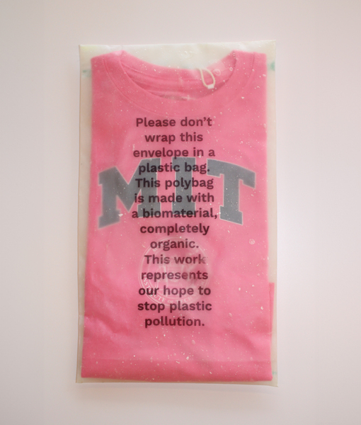 A folded, pink MIT t-shirt is wrapped in translucent packaging. The wrapping reads "Please don't wrap this envelope in a plastic bag. This polybag is made with a biomaterial, completely organic. This work represents our hope to stop plastic pollution."