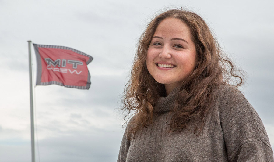 MIT Senior Tatum Wilhem is pictured close up, outdoors against a gray sky, with an MIT Crew flag flying in the background.