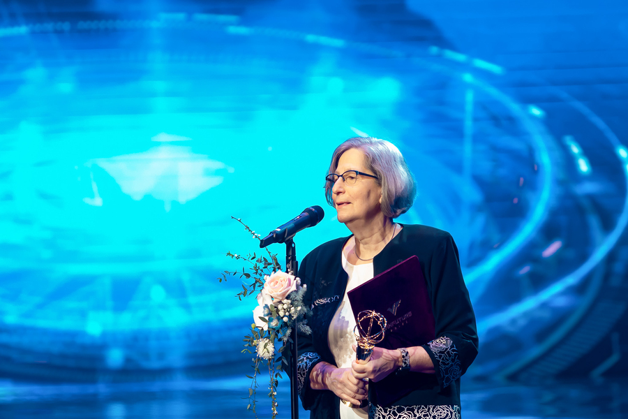 Susan Solomon stands on a stage, speaking into a microphone with a floral corsage on its stand. She is holding a gold trophy in one hand.