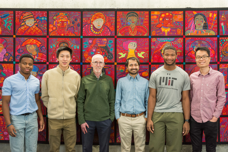 Six people stand facing the camera against a red, quilted mural with faces on it
