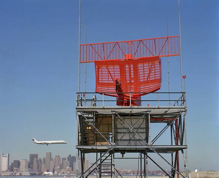 Exterior photo of a large airport radar antenna. In the background is a low-flying commercial aircraft with the skyline of Boston behind that.