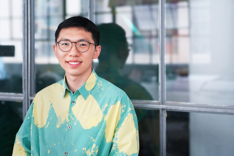 Chen Chu, male student with short hair wearing glasses and a colorful shirt, sits in front of a glass wall, smiling.