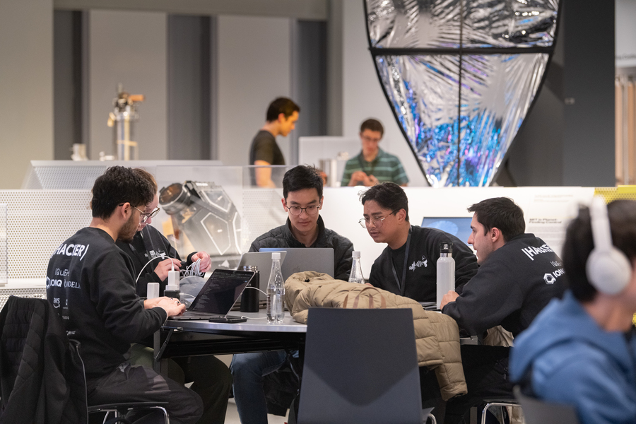 Five students gather around a table strewn with laptops, chargers, and other equipment.
