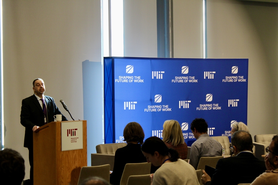 Daron Acemoglu speaks in front of an audience from a lectern next to a sign with the MIT logo and "Shaping the Future of Work."