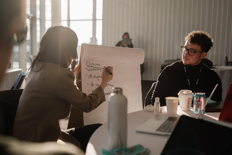 Two people sitting down, with one whose back is to the camera writing on a large notepad.