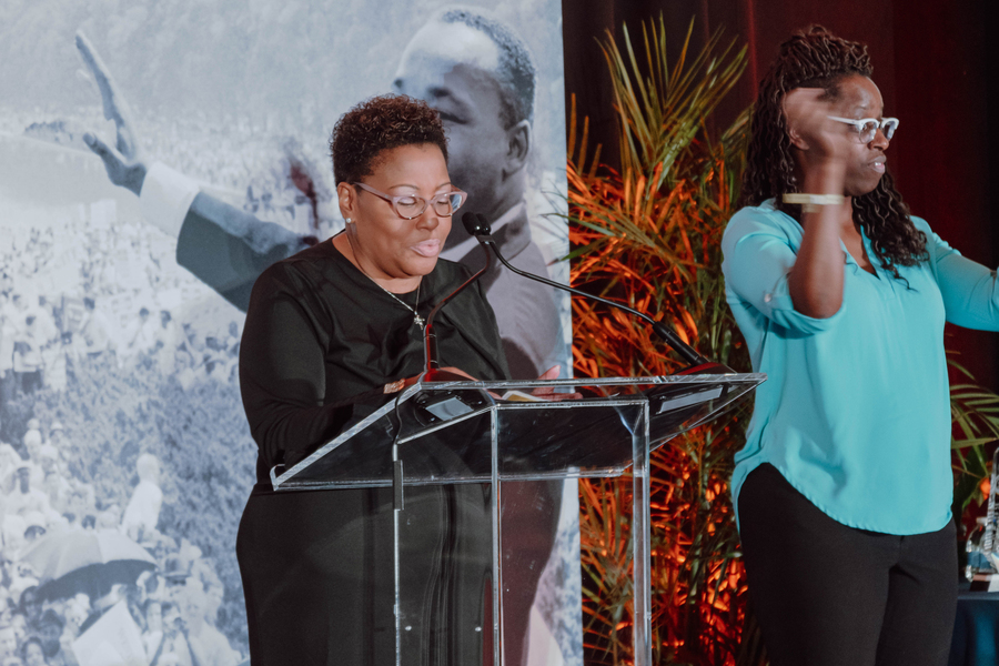 DiOnetta Jones Crayton stands on stage with an interpreter. A greyscale photo ofMartin Luther King Jr. at the 1963 March on Washington is in the background.