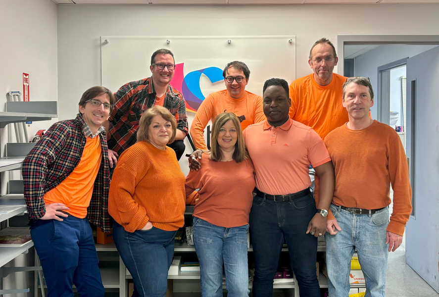 Eight Copytech team members wear orange shirts and pose for a photo in their office.