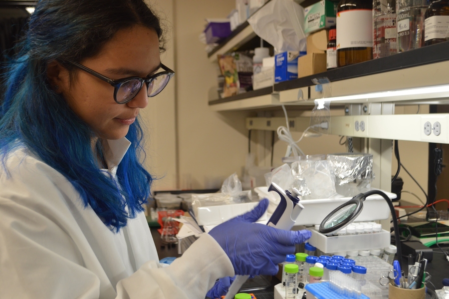 Iraira Rivera Rojas, sporting long blue hair, glasses, and a lab coat, holds a measurement instrument in a lab.