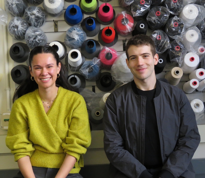 A woman and a man sit in front of colorful spools of yarn, smiling for the camera. 
