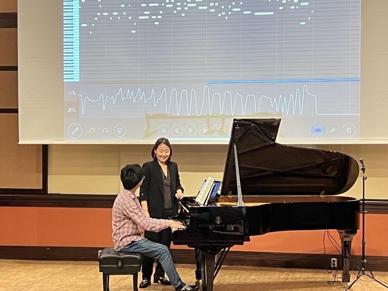 William Wang plays a piano while Mi-Eun Kim watches; behind them, data are projected on a screen.