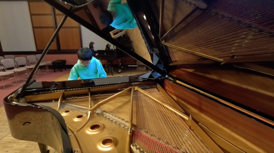 Holden Mui, looking down, plays the piano. He is seen from the perspective of an open piano case in the foreground