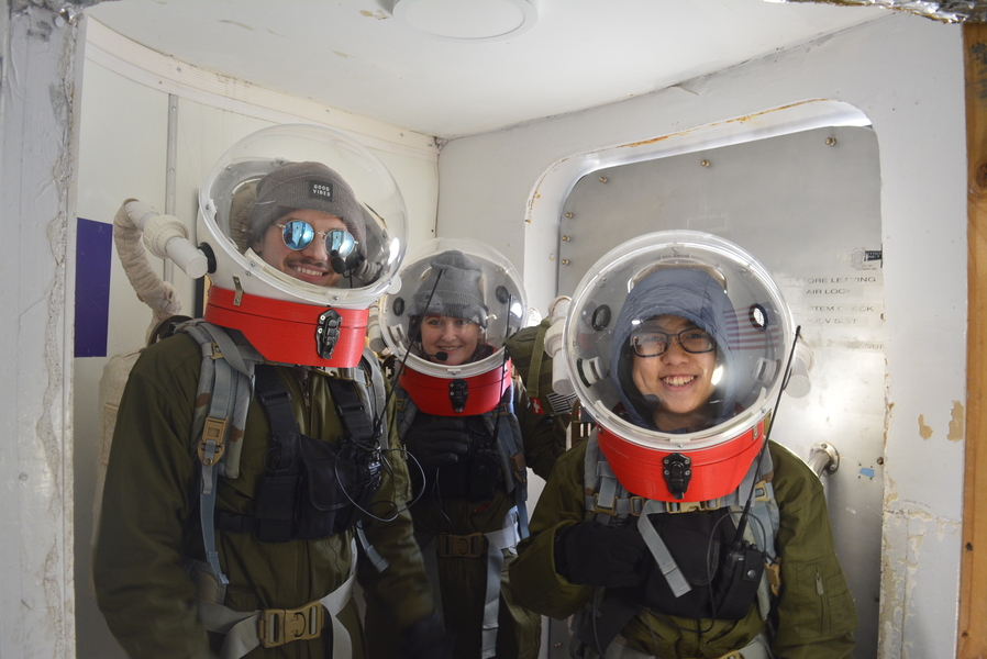 Benjamin Kazimer, Madelyn Hoying, and Wing Lam (Nicole) Chan, all wearing EVA suits, smile through their bubble helmets near the hatch door inside a research facility.