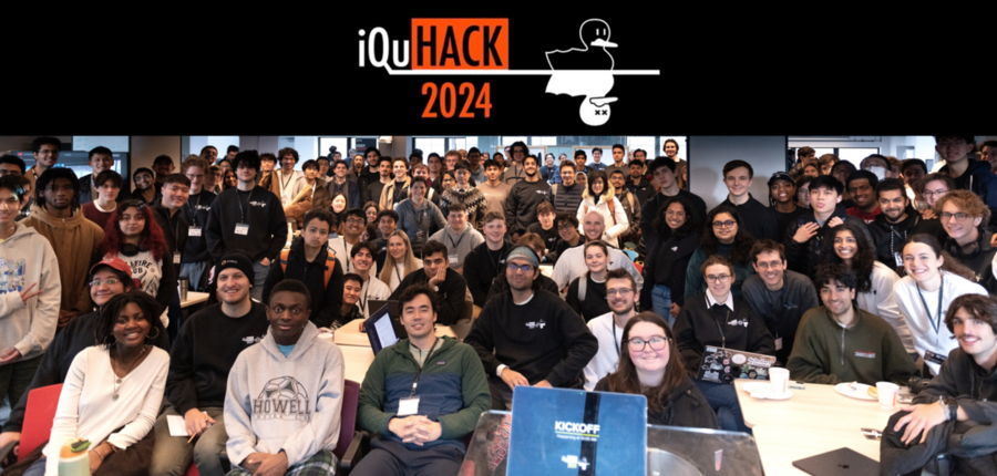 Perhaps 150 people pose for a photo in a large classroom. About a third are seated in the foreground and the rest stand. At top is a banner that reads iQuHACK 2024, with the image of two nearly identical ducks, one alive and one shown as dead with X's for eyes