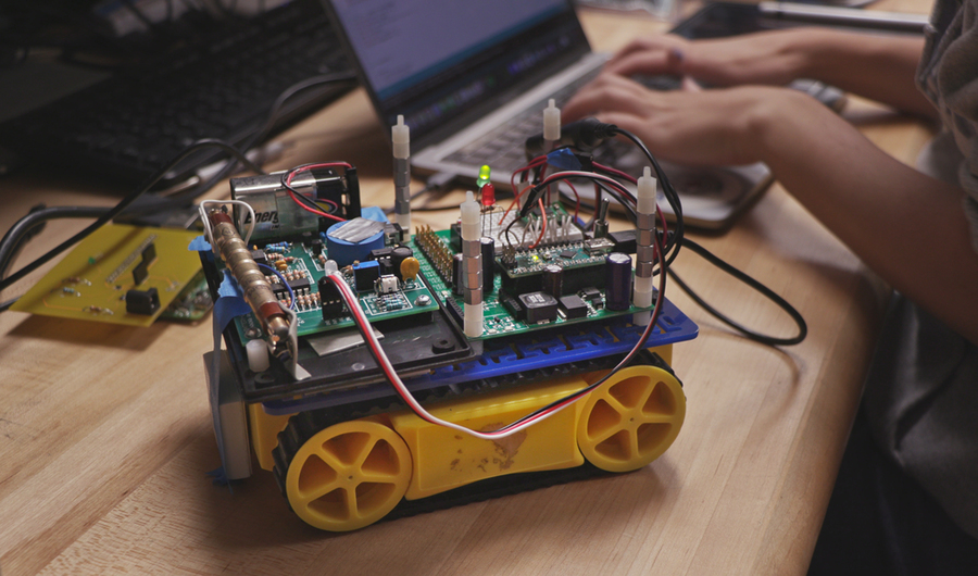 A small, rectangular robot sits on a table top; a student's hands are seen typing on a laptop in the background