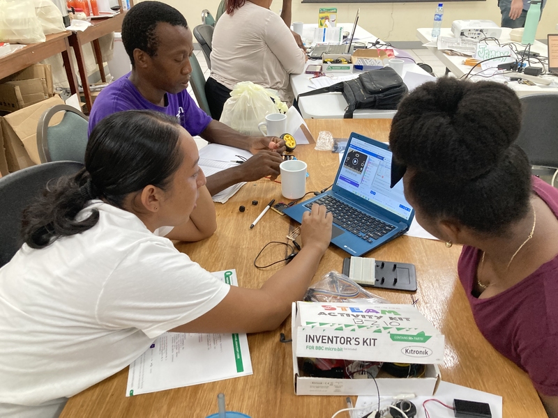 Three people sit around a cluttered table, looking at a laptop