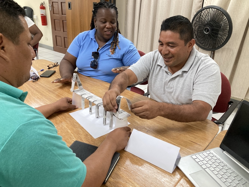 Three people sit at a table creating a structure made out of pieces of newspaper and two plastic cups