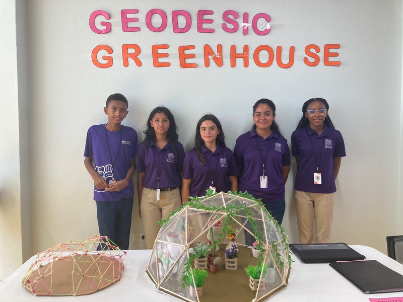Five children wearing purple shirts stand against a wall displaying the words “Geodesic Greenhouse.” Two geodesic models are on a table in front of them.