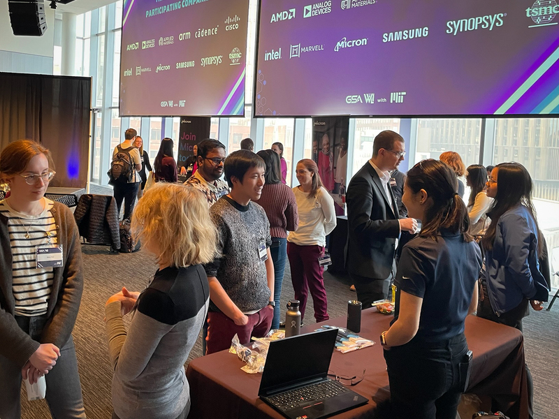 About two dozen students and company representatives stand chatting among tables in a conference room with floor-to-ceiling windows. Two large hanging screens display various company logos