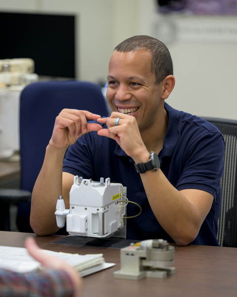Christopher Williams sits at a table with a small cube-like piece of equipment before him. He smiles as he gestures with both hands