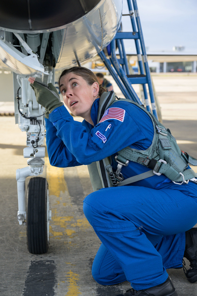 Christina Birch, wearing a blue flightsuit, crouches while holding open a panel underneath a fighter jet at an airfield 