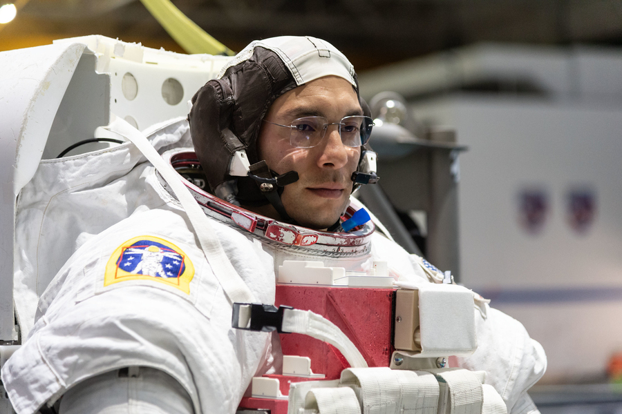 Closeup of Marcos Berrios wearing a white spacesuit in an indoor facility. His head is covered with a cap and audio equipment but his helmet is off