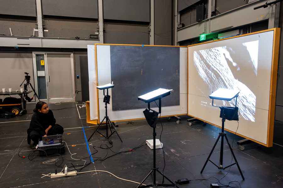 In a room with lots of open space, a person crouches beside a projector that shines images of artwork with white and black design onto a screen. In between her and the screen are three lights mounted on tripods.