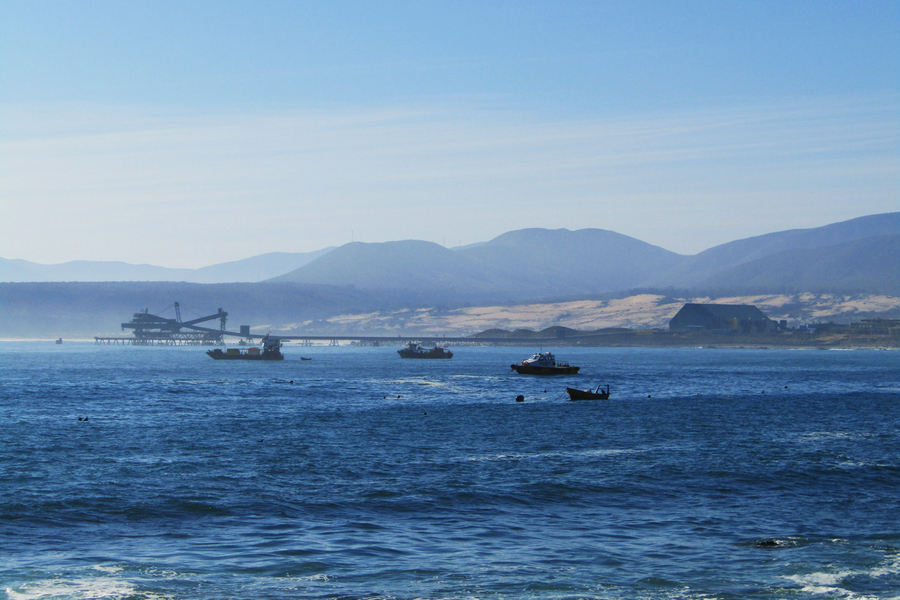 Blue ocean with four boats in the foreground, and crane-like machinery at a port, mountains, and a building that is part of a desalination plant in the background.