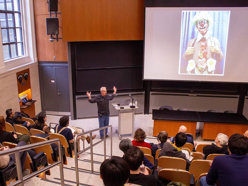 Master Bladesmith Bob Kramer speaks in front of an audience in a classroom with stadium seating. Behind him on a screen is a picture of Kramer as a circus clown.