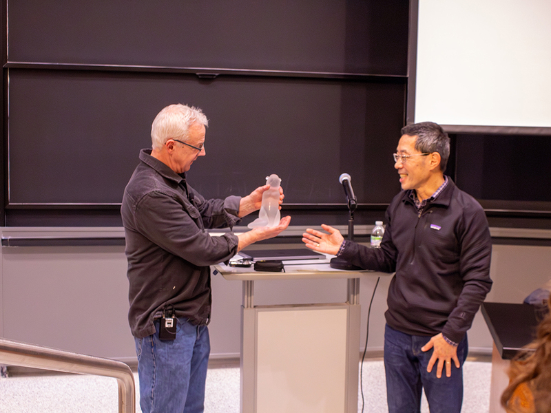 Two men flank a lectern in front of a classroom. One is presenting a glass beaver sculpture to the other. 