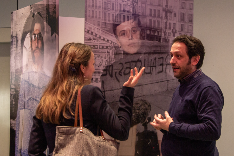 Pierre-Jérôme Adjedj, seen in profile, converses with a woman with long hair whose back is to the camera in front of two poster-sized photo collages of men in front of buildings