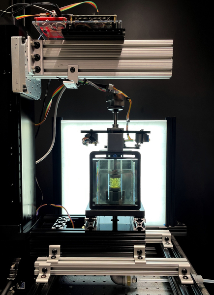 In the lab, the prototype scanner is made of metal and wires, with a sturdy clear box inside. Inside the box is a glass tube with yellow material inside. A lightbox is in the background, providing light.