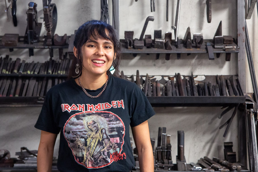 Amber Velez, wearing an Iron Maiden T-shirt stands in front of a tall shelf of metal-working tools.