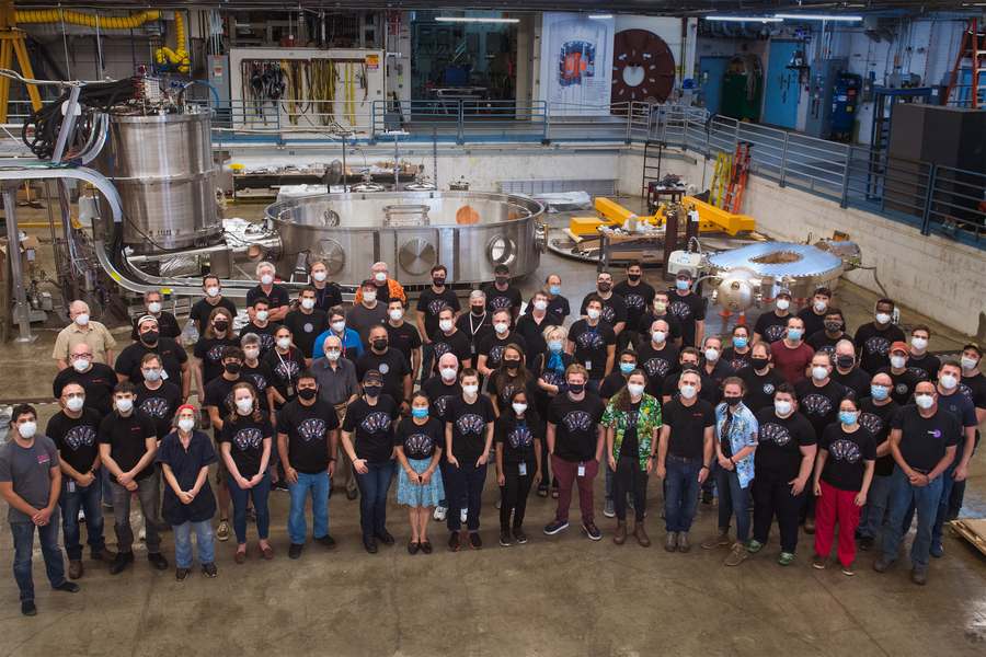 Group of about 70 people wearing face masks stand in front of the cryostat container 