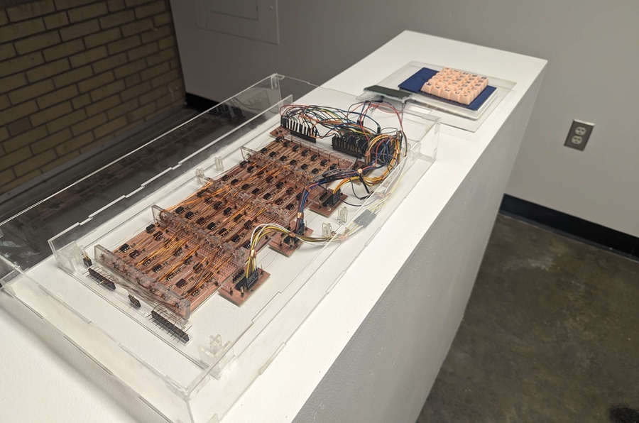 A core rope memory prototype, which is a flat array of circuits and wires, sits on a white pedestal in a gallery.