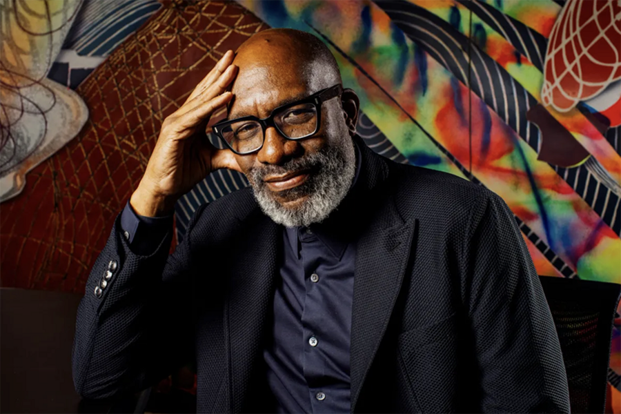 Portrait photo of Larry Sass, a Black man with dark-rimmed glasses and a beard, seated before a colorful mural