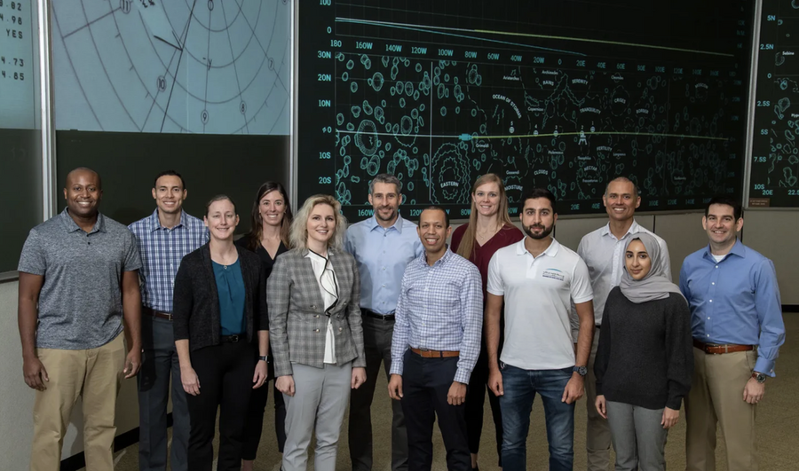 A dozen casually dressed people stand in a room with scientific information on a board behind them